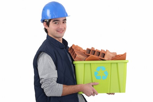 A young man wearing a blue safety helmet and a navy vest over a grey long-sleeve shirt is holding a bright green recycling bin with a blue recycling symbol on the side, filled with red bricks and construction debris. He is smiling and positioned against a plain white background. The bin appears to be made of sturdy plastic with a smooth finish. The bricks inside are irregularly shaped, with some showing stacked layers and others partially broken. The scene emphasizes waste collection and disposal, relevant to rubbish removal services such as those offered by Business Waste Removal Plaistow, serving areas in and around the Plaistow postcode district in East London. The lighting is even, highlighting the textures and colors of the bricks and the smooth surface of the bin, creating a professional and clear representation of waste disposal related to construction or renovation debris.