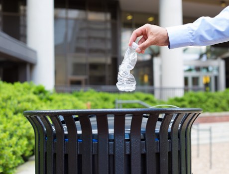 A person's hand, wearing a light blue shirt sleeve, is seen dropping a crumpled transparent plastic water bottle into a black, slatted outdoor rubbish bin situated on a paved area. The background includes modern office or commercial building structures with glass windows and metal framing, along with green shrubbery bordering the scene. This outdoor setting highlights waste disposal in a professional or urban environment near Plaistow, with the bin positioned on a concrete or stone surface. The plastic bottle, visibly crumpled, is in mid-air as it is about to be deposited into the bin, reflecting typical rubbish collection behaviour. Business Waste Removal Plaistow offers services that include the collection and disposal of such waste materials, consistent with local urban waste management needs. The scene emphasizes the importance of proper waste disposal in public or commercial spaces, supporting effective rubbish removal practices in the area.