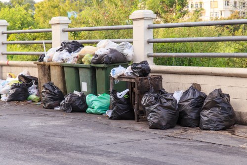 A collection of black, green, and white rubbish bags are piled along the edge of a paved roadway adjacent to a concrete bridge with metal railings. Some bags rest on the pavement while others are stacked on top of a wooden bench-like structure mounted against the railing. The bags appear to contain general waste and are of various sizes and shapes, with some slightly torn or bulging. In the background, greenery and trees are visible, suggesting a park or riverbank setting, with hints of buildings in the distance. The overall scene depicts a typical outdoor refuse collection area in the vicinity of Plaistow, with the bags awaiting collection by waste removal services. Business Waste Removal Plaistow operates in this area to manage such rubbish accumulation efficiently and professionally.