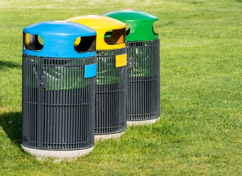 Three cylindrical outdoor rubbish bins are positioned on a grassy area, each with a black, vertical slatted design and a rounded lid in bright blue, yellow, and green colors. The blue-lid bin is in the foreground, followed by the yellow and green-lidded bins, which are slightly behind and to the right. The bins appear to be made of durable plastic, with textured finishes on their surfaces, and are situated on a well-maintained lawn. The clear sky suggests daytime lighting, with natural sunlight illuminating the scene. The placement of the bins and their vibrant colors indicate they are designated for different types of waste, aligning with local rubbish management practices. These bins are located in an outdoor setting, possibly on a residential driveway or communal area, representative of waste disposal services in Plaistow or similar neighbourhoods. Business Waste Removal Plaistow offers tailored rubbish collection solutions, as reflected in the organized setup of waste receptacles.