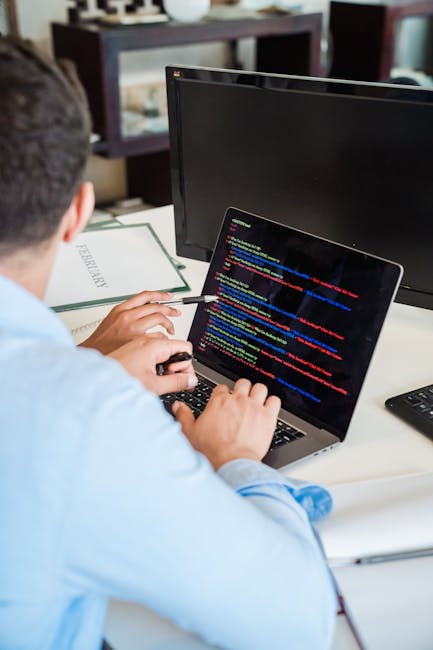 A person with short dark hair, wearing a light blue shirt, is sitting at a desk using a laptop with a black screen displaying colorful lines of programming code or digital text. The individual is posi