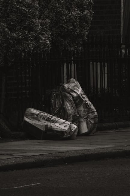 An overflowing collection of various waste materials, including black, black-and-blue, and red waste bins filled with mixed rubbish, opened and overfilled with cardboard boxes, plastic bags, paper, an