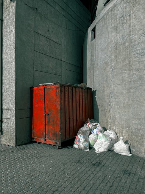 A large, weathered red metal skip with visible rust and scratches is positioned against a dark green wall on the left side and a light grey concrete wall on the right side of the image. The skip is si